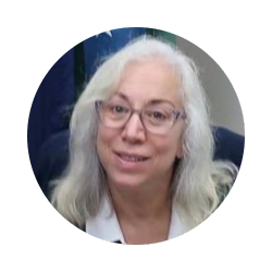 A woman with light hair wearing glasses and a white collared shirt is photographed against a blue and white background with a flag behind her.